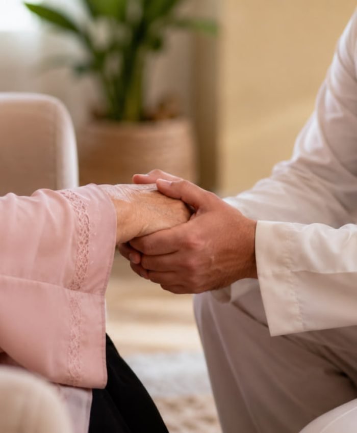 An adult Muslim son holding his elderly mother's hand with deep respect and care in a warm home environment.